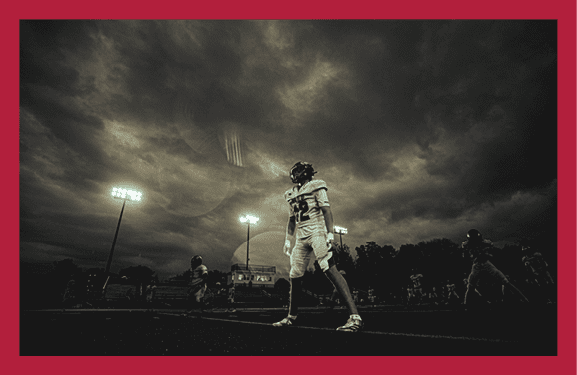A person wearing a football helmet and uniform looks upward against a dramatic, cloudy sky with a dark and moody atmosphere. Description generated by AI