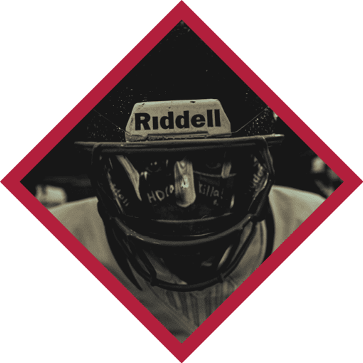Person wearing a Riddell football helmet and jersey is looking downward in a close-up shot framed by a red diamond border. Description generated by AI