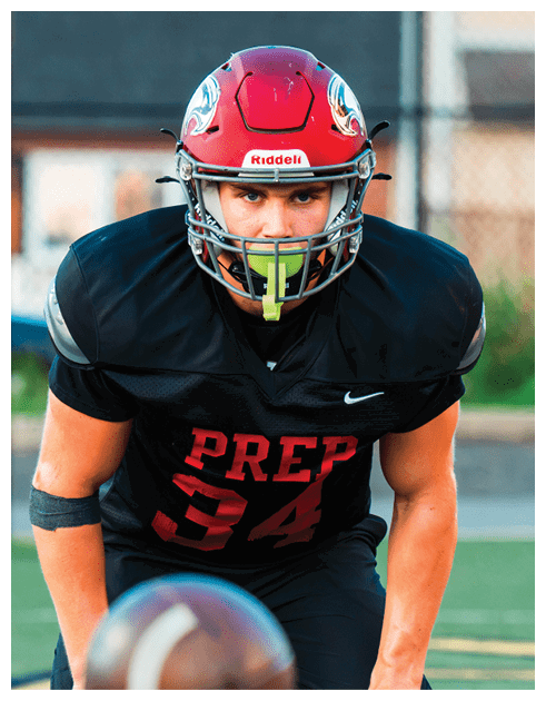 A person wearing a black football uniform with the word PREP and the number 34 in red is crouched on a field with a football in the foreground. Description generated by AI