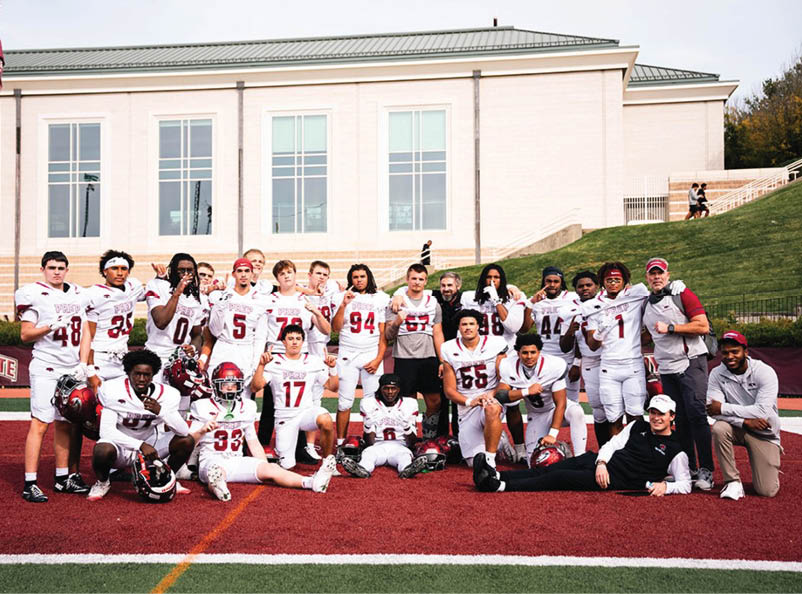 A group of people in football uniforms and casual clothing pose together on a sports field in front of a building. Description generated by AI