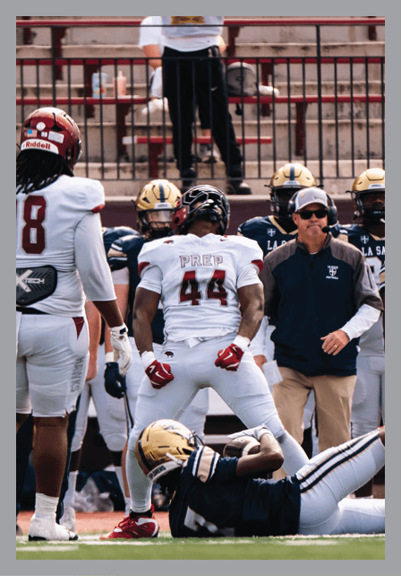A group of football players and a coach stand on the field with one player lying on the ground during a game. Description generated by AI