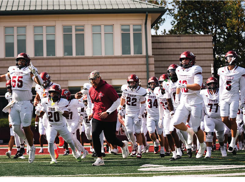A group of people in white football uniforms and helmets are running onto a field alongside a person in a red jacket and cap. Description generated by AI
