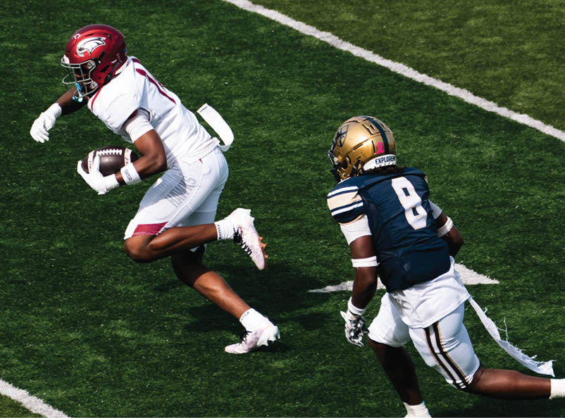 A person in a white football uniform with a red helmet is running with the ball while another person in a navy blue and white football uniform with a gold helmet is chasing behind on a green field. Description generated by AI