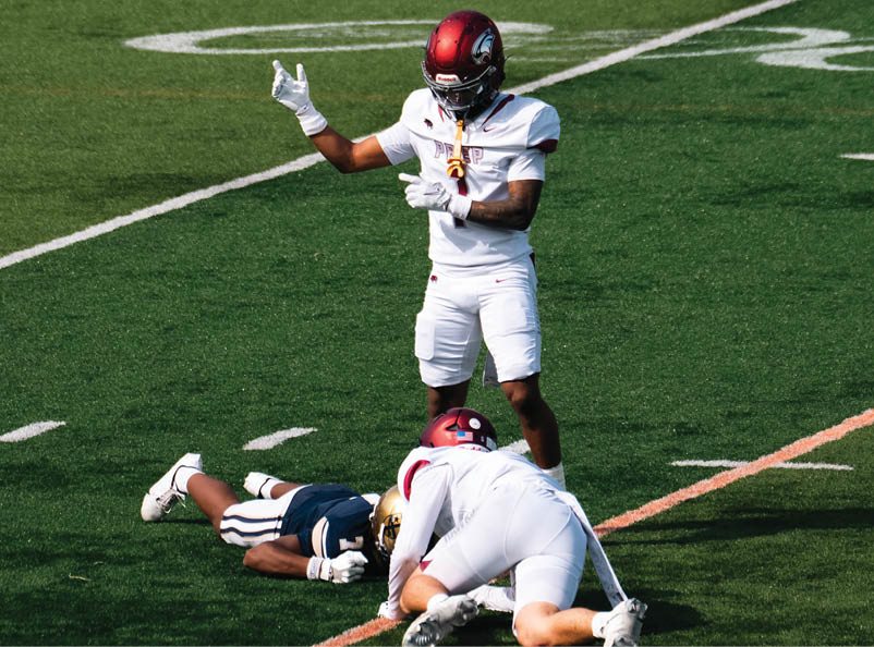 A person in a white football uniform stands on a field gesturing with one hand while two other people in football uniforms are on the ground nearby. Description generated by AI