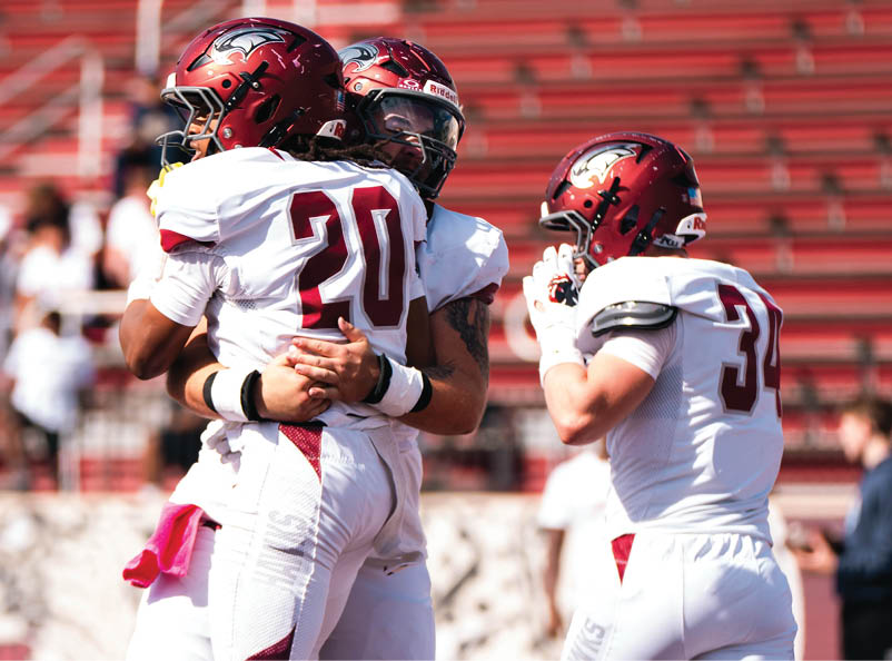 Three people in football uniforms and helmets are celebrating on a field with red bleachers in the background. Description generated by AI