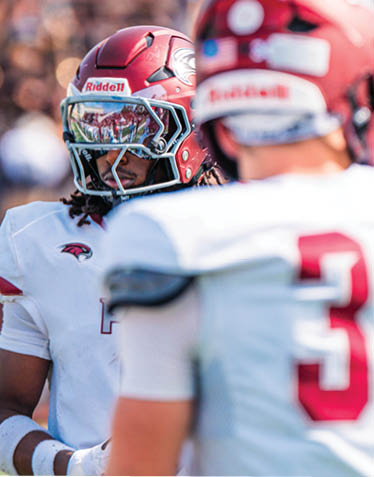 Two people wearing red helmets and white football uniforms stand on a field with blurred spectators in the background. Description generated by AI