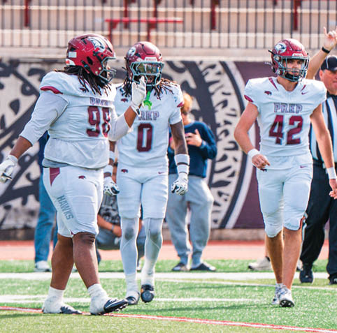 Three people in football uniforms and helmets stand on a field with one person gesturing with their hand near their mouth. Description generated by AI