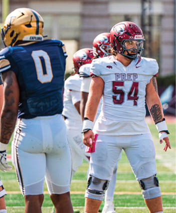 A group of people in football uniforms stand on a field with one person in white and red gear positioned behind a football ready for play. Description generated by AI