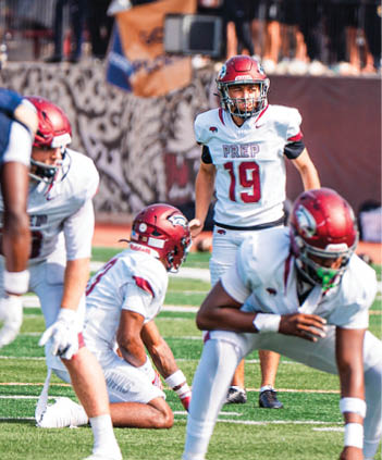 Four people in football uniforms and helmets are positioned on a field, with one person kneeling and the others standing or crouching, appearing ready for a play. Description generated by AI