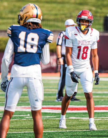Two people in football uniforms stand on a field with one wearing a white uniform and red helmet and the other in a blue uniform and gold helmet. Description generated by AI