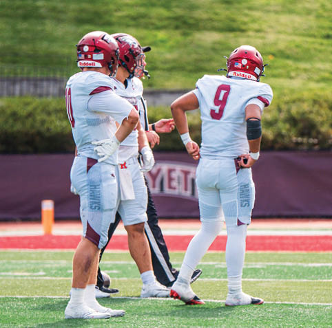 Three people wearing white football uniforms and red helmets stand on a field with green grass and a maroon banner in the background. Description generated by AI