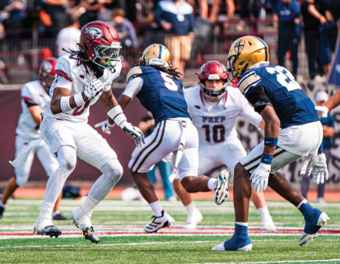 Four people in football uniforms are actively engaged on a football field with a blurred crowd in the background. Description generated by AI