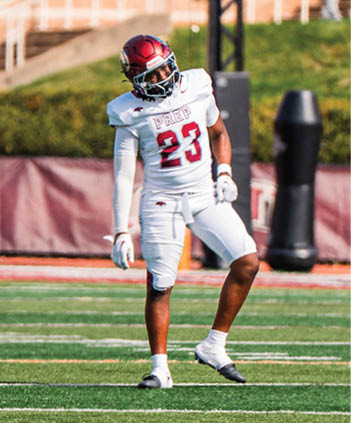 A person wearing a white football uniform with the number 23 is standing on a football field with a stadium and another person in the background. Description generated by AI