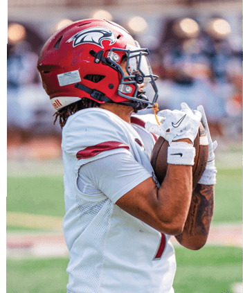 A person wearing a red football helmet and white uniform is holding a football close to their chest on a grassy field with blurred spectators in the background. Description generated by AI