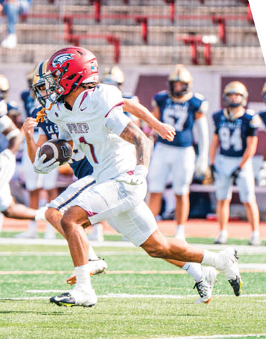 A person in a white football uniform with a red helmet is running on a field while holding a football, pursued by several people in dark uniforms. Description generated by AI