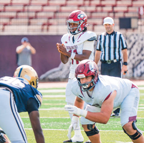 A person in a white football uniform and red helmet is preparing to throw a football while another person in a white uniform is crouched in a blocking stance facing an opponent in a dark blue uniform on a football field with a referee in the background. Description generated by AI