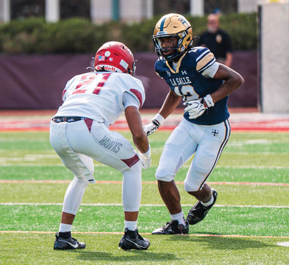 Two people in football uniforms are facing each other on a field, with one person in a blue and gold uniform and the other in a white and red uniform. Description generated by AI