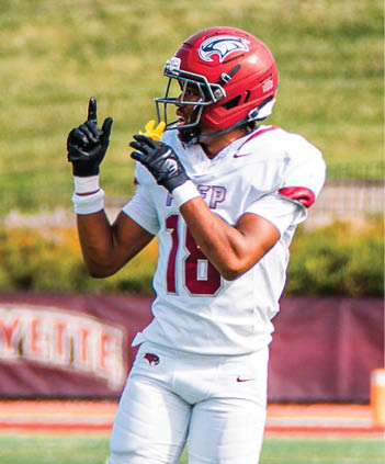 A person wearing a red football helmet and white uniform with the number 16 is standing on a football field pointing upwards with both hands. Description generated by AI