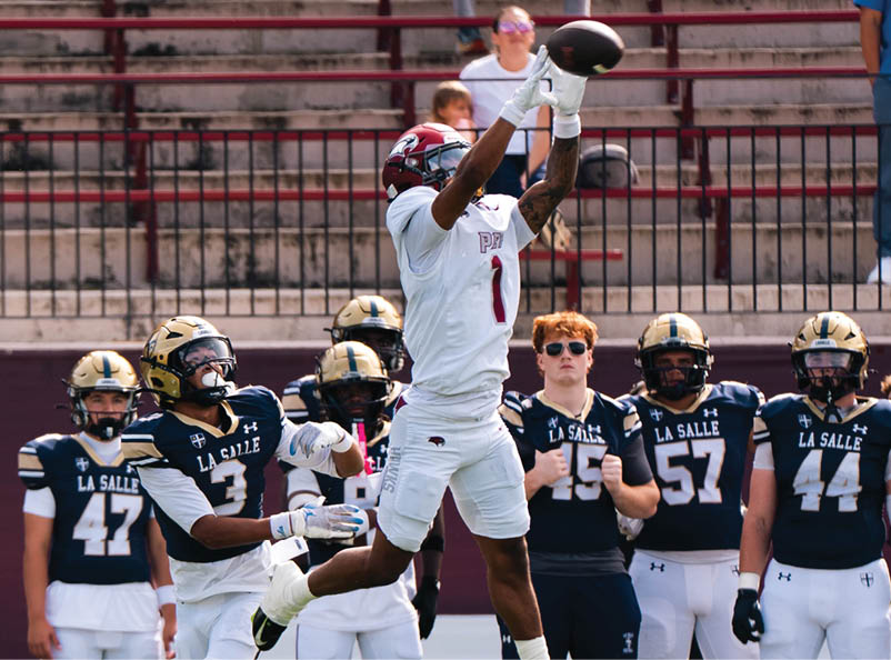A person in white sports gear jumps to catch a football while several people in dark blue uniforms watch on a field with empty bleachers in the background. Description generated by AI