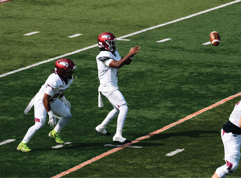 Three people in white football uniforms and red helmets are on a green field with one person catching a football. Description generated by AI