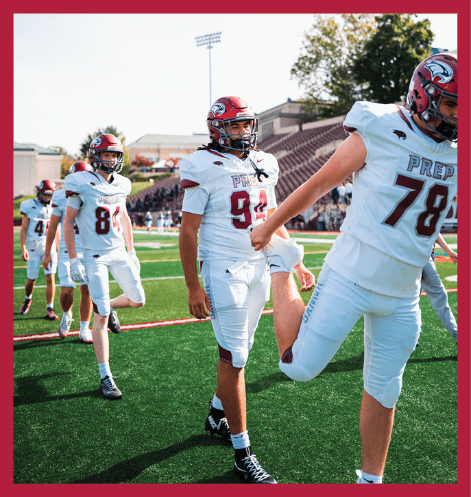 Several people in white football uniforms and red helmets are walking on a green field with stadium seating in the background. Description generated by AI