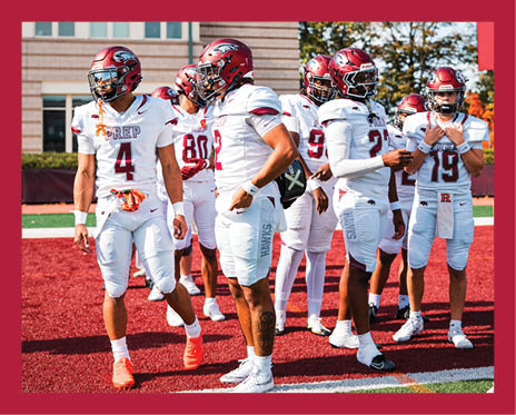 A group of people wearing white football uniforms and helmets are standing on a red and green field, appearing to prepare for a game or practice. Description generated by AI