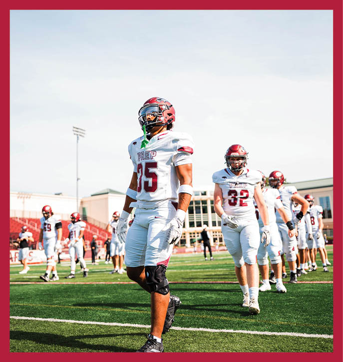 Several people in football uniforms and helmets are standing and walking on a green field with a stadium and clear sky in the background. Description generated by AI