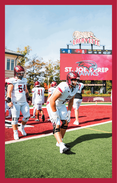 Several people in football uniforms and helmets are standing and preparing on a field near a sign that reads “ST. JOE HAWKS." Description generated by AI