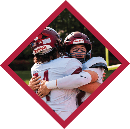 Two people in football uniforms and helmets are embracing, with one person showing eye black and a visible expression of emotion. Description generated by AI