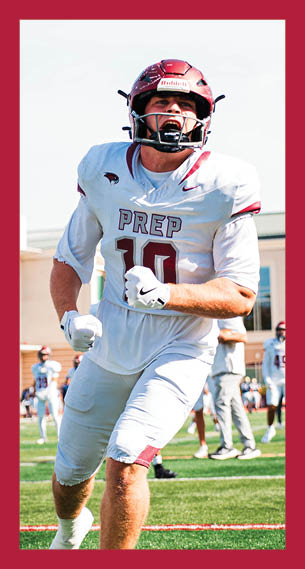 A person in a white football uniform and helmet is running on a green field with another person standing behind wearing a similar uniform and helmet, with a building and other players in the background. Description generated by AI
