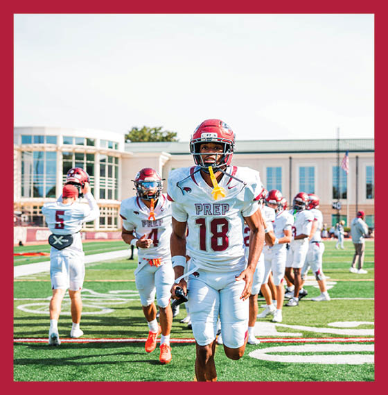 People in football uniforms are running and walking on a green field with yard lines and a stadium in the background. Description generated by AI