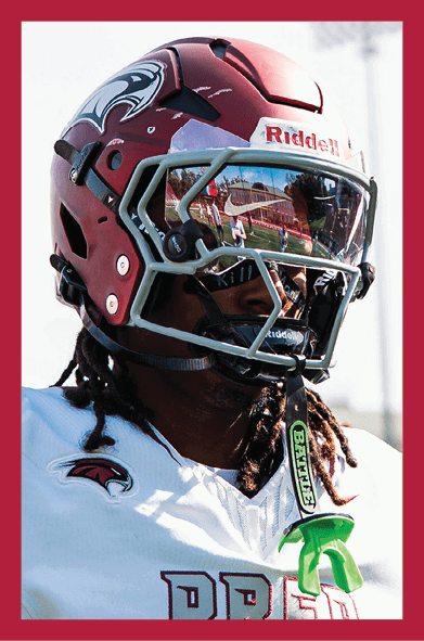 A person wearing a maroon football helmet with a reflective visor and a white sports jersey with a bird logo is shown in close-up. Description generated by AI