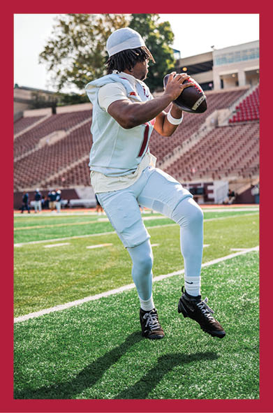 A person wearing a white football uniform and a white head covering is preparing to throw a football on a green field with empty stadium seats in the background. Description generated by AI