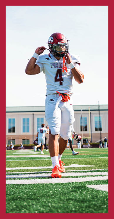A person in a football uniform with a red helmet and orange cleats is standing on a green field with a building in the background. Description generated by AI