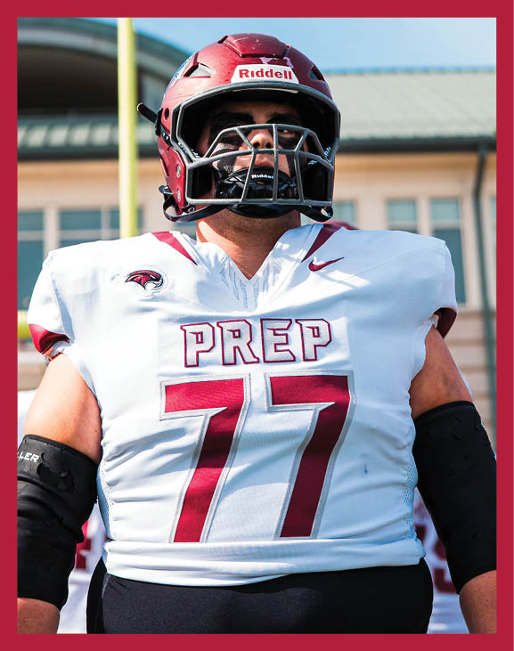 A person wearing a white football uniform with the number 77 and the word PREP stands on a football field in front of a goalpost and a building under a clear blue sky. Description generated by AI