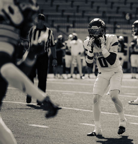 A person in a football uniform and helmet is preparing to catch a ball on a field while a referee and other players stand in the background. Description generated by AI