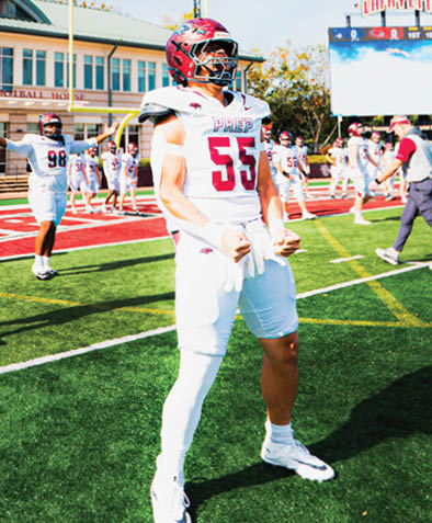 A person in a football uniform with the number 55 is standing on a football field with other players and staff in the background near a scoreboard and goalpost. Description generated by AI
