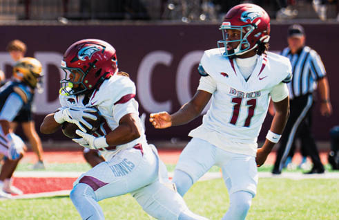 Two people in white football uniforms with red helmets are playing on a field, with one holding the ball and the other appearing to prepare to block or assist. Description generated by AI