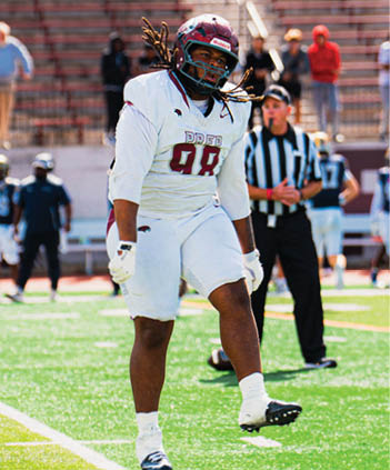 A person wearing a white football uniform with the number 98 and a helmet is standing on a football field with a referee and other people in the background. Description generated by AI