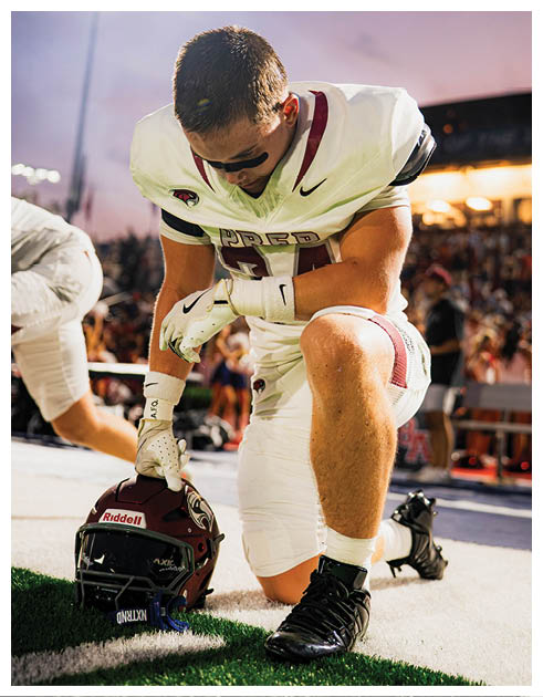 A person in a football uniform kneels on one knee on the field while holding a football helmet with one hand. Description generated by AI