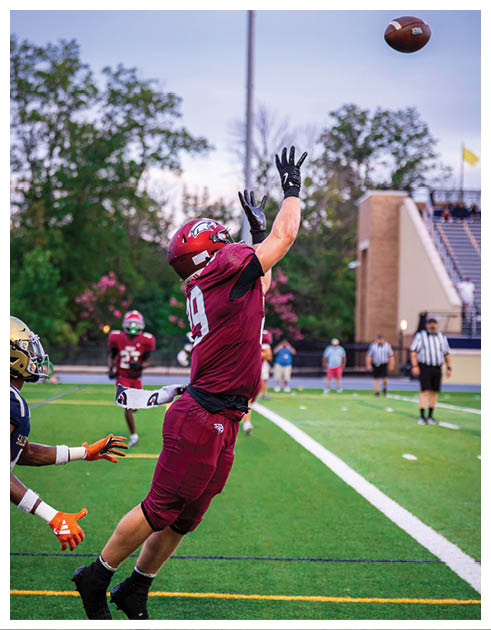 A person in a maroon football uniform is jumping with arms raised on a green field while another person in a dark uniform reaches toward them. Description generated by AI