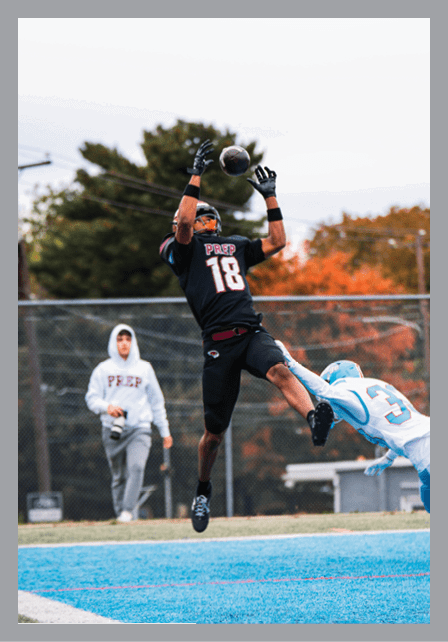 A person in a black sports uniform with the number 18 is jumping to catch a football while another person in a light blue and white uniform attempts to block them on a field with a fence and trees in the background. Description generated by AI