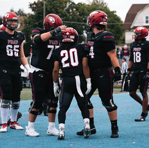 A group of people wearing black football uniforms with red helmets are gathered on a blue sports field during a game. Description generated by AI