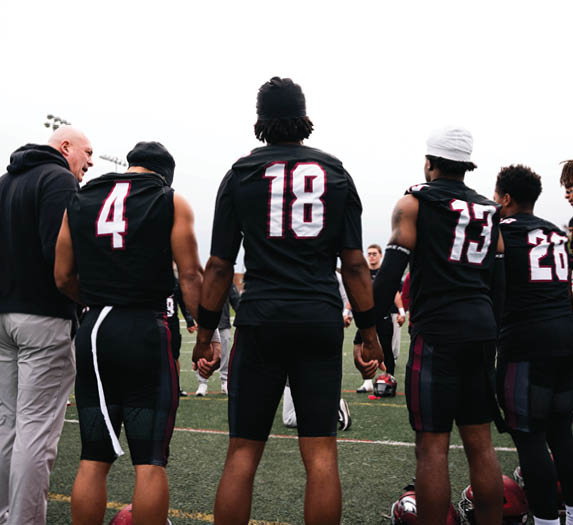 People wearing black sports uniforms stand on a field with red helmets placed on the ground in front of them. Description generated by AI