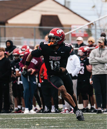 A person wearing a red helmet and black football uniform with the word “PREP" is running on a football field with a group of people standing in the background. Description generated by AI