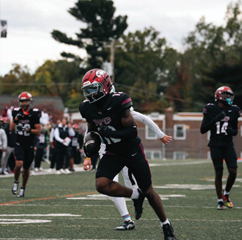 A person wearing a red helmet and black football uniform is running with a football on a field while other players and spectators are visible in the background. Description generated by AI