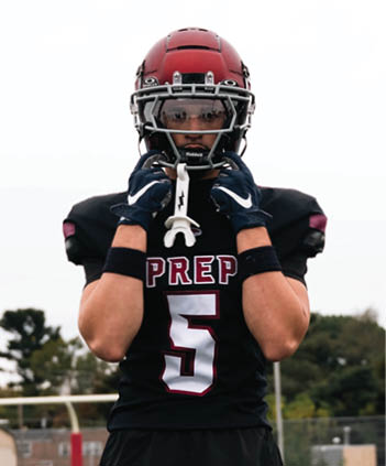 A person wearing a black football uniform with the number five and a red helmet stands on a football field with other players and goalposts visible in the background. Description generated by AI