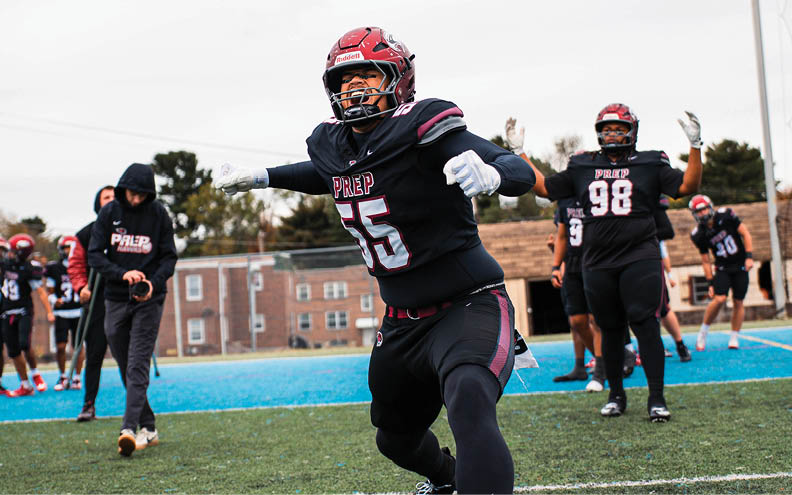 A person in a football uniform with the number 55 is running energetically on a field while other players and a coach stand in the background. Description generated by AI