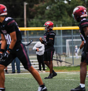 Three people in football uniforms and helmets stand on a field with a fence and trees in the background. Description generated by AI