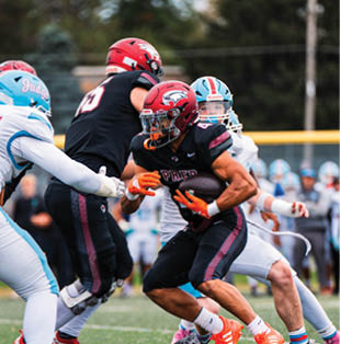 An adult wearing a black and maroon football uniform is holding a football tightly while another adult in a white uniform reaches out behind them on a football field. Description generated by AI